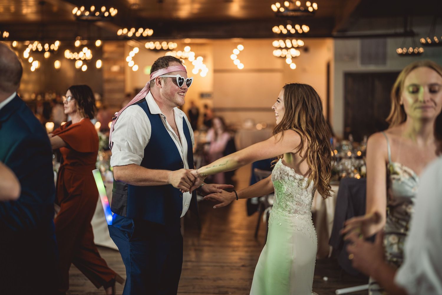 Guests dancing joyfully at a lively wedding reception with warm string lights in the background.