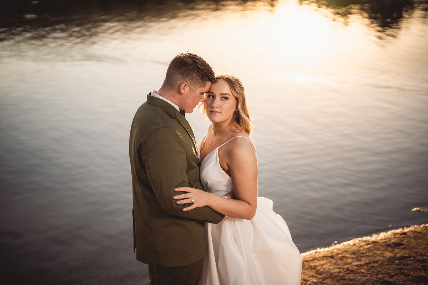 Couple embracing by a golden sunset lake, bride in white gown and groom in olive suit, romantic outdoor wedding photo.