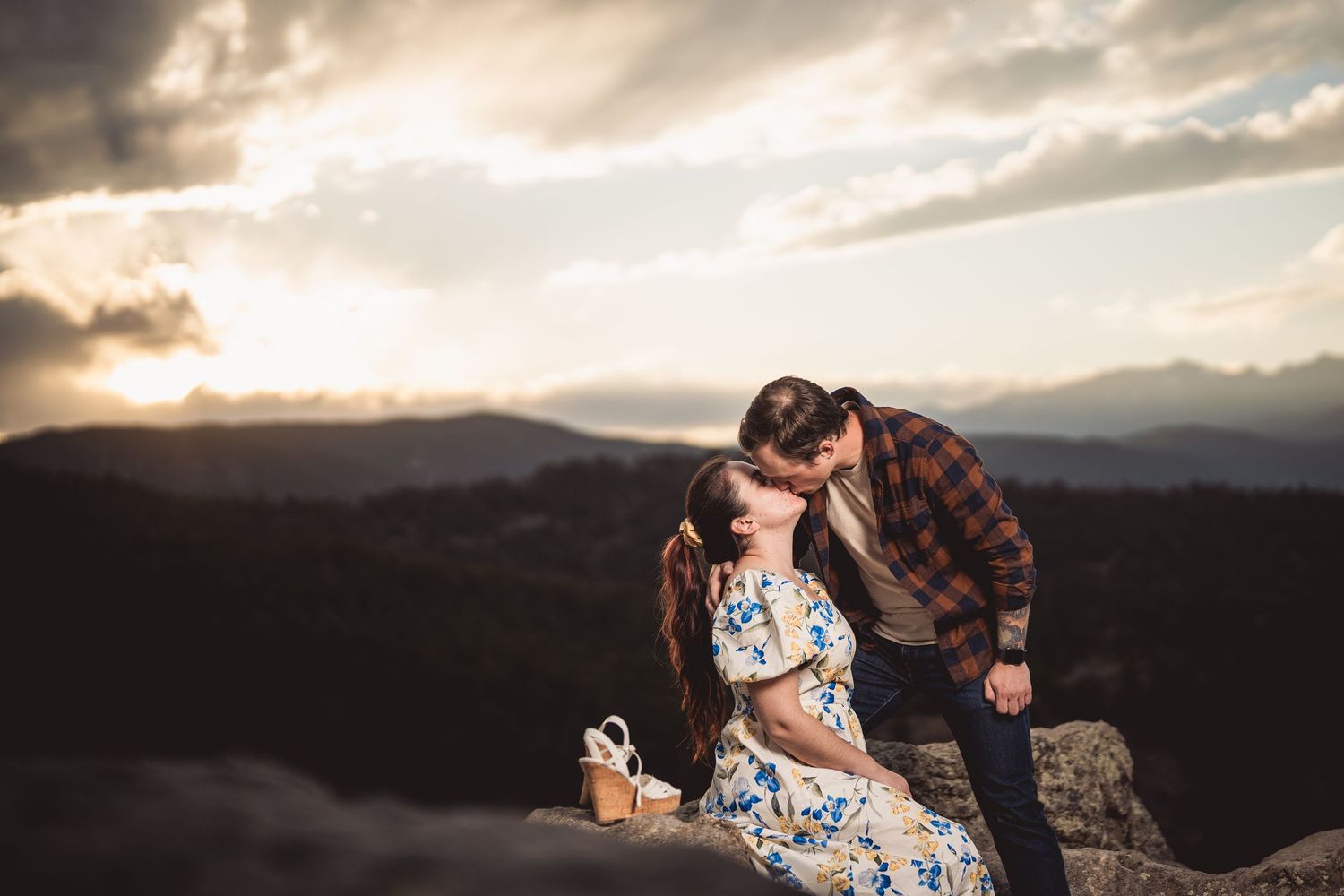 Couple sharing a romantic kiss on a mountain overlook at sunset with dramatic clouds and scenic valley views.