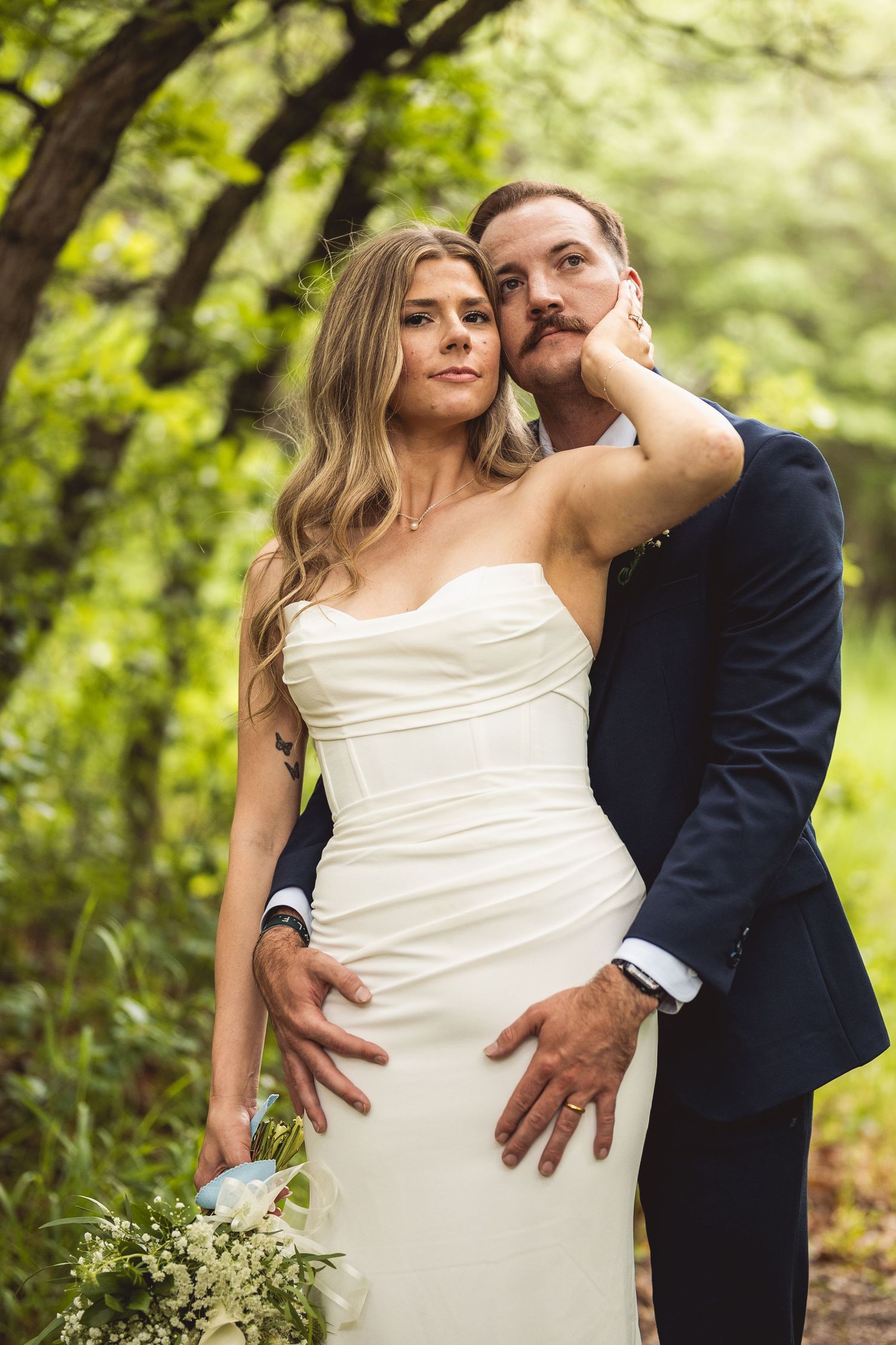 A bride in a white strapless gown and groom in navy suit embrace romantically outdoors amid lush green trees.