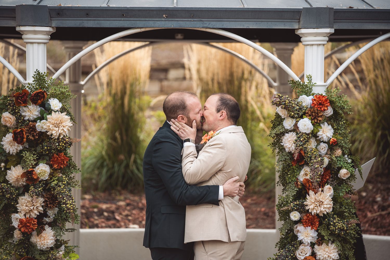 Two grooms share their first kiss under a floral arch adorned with red and white flowers at an outdoor wedding ceremony.