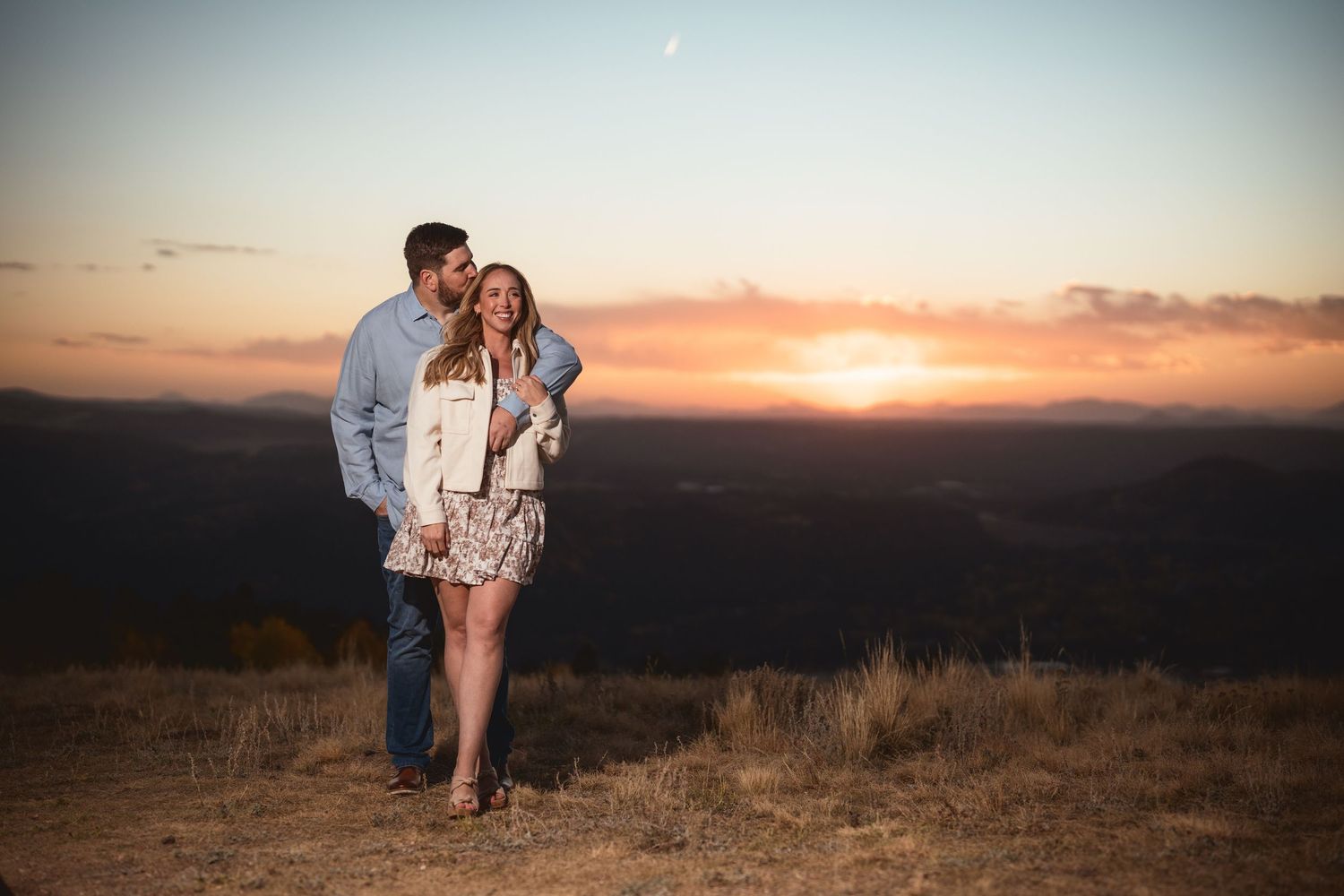 Couple embracing at sunset on a mountain overlook, warm golden sky behind them.