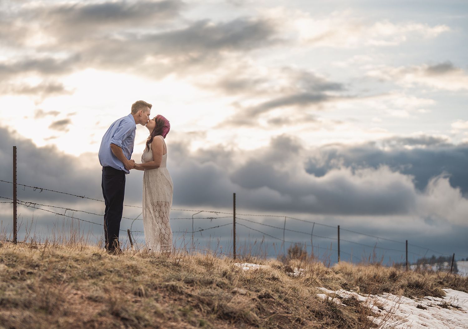A couple shares a romantic kiss at sunset on a hilltop, with dramatic storm clouds filling the sky behind them.