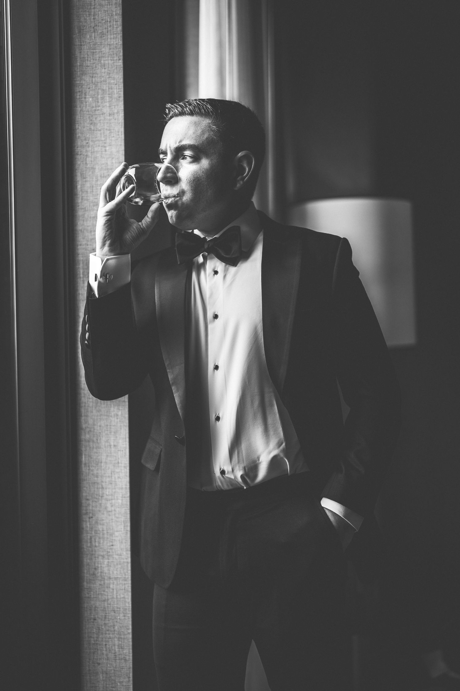 A dapper man in a tuxedo with bow tie sips a drink by a window in a classic black and white portrait.