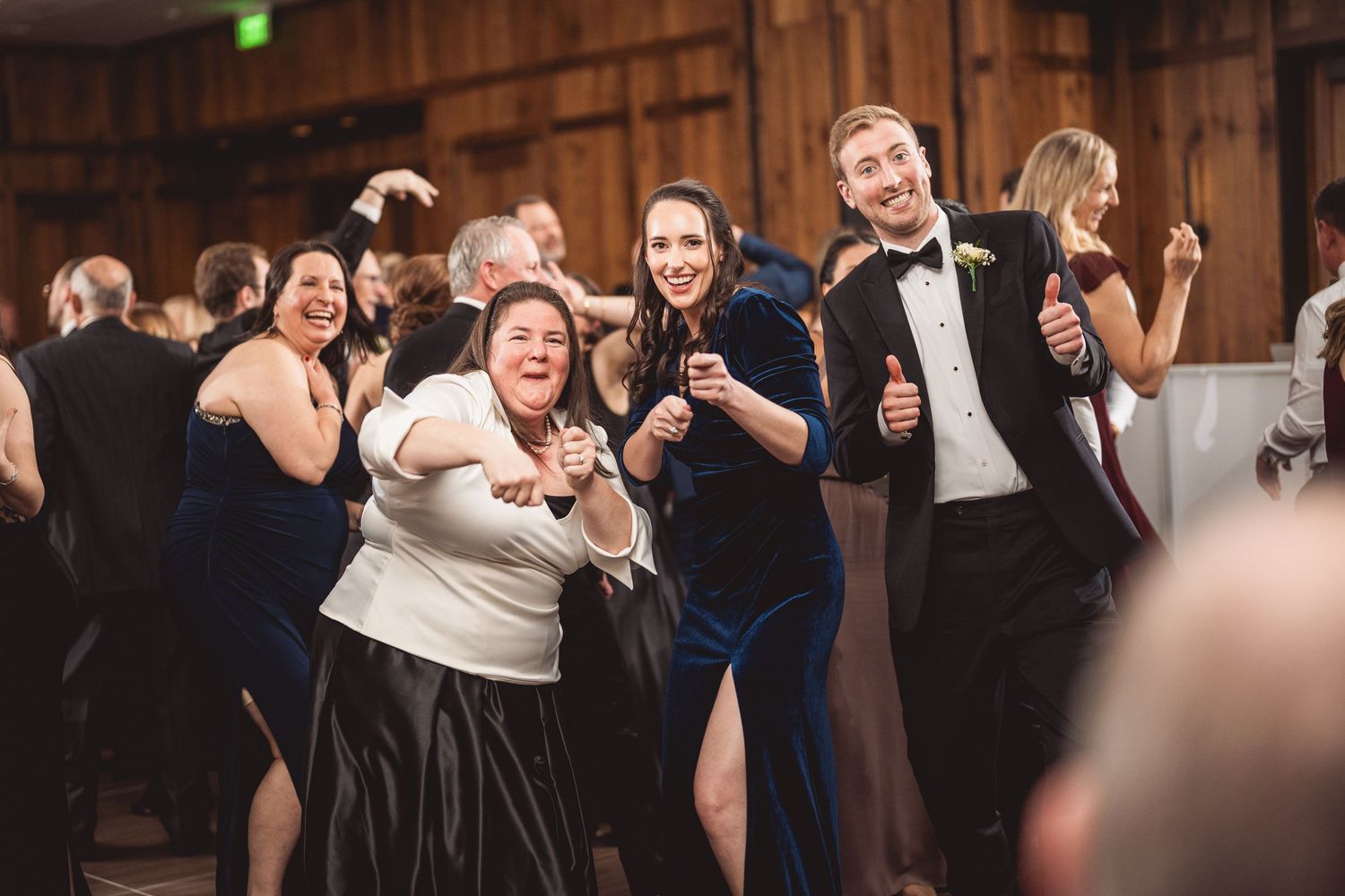 Wedding guests dance and celebrate on a wooden dance floor at an indoor reception venue.