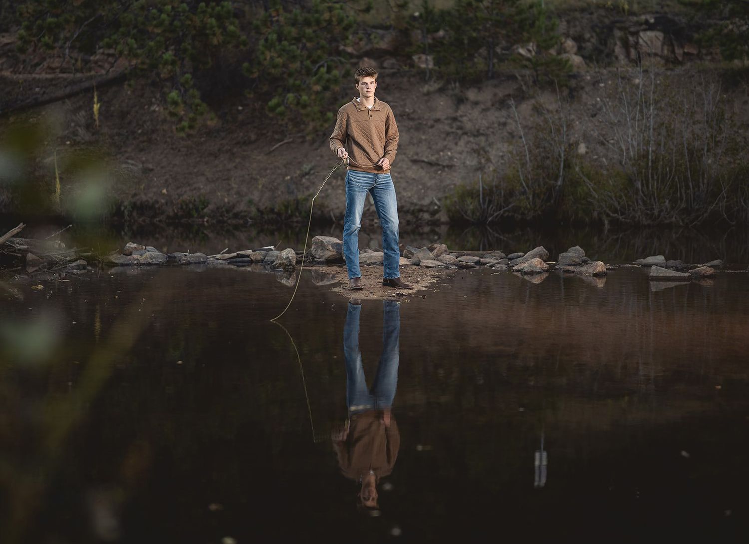 Young man standing on rocks reflected in still water holding a fishing rod in moody outdoor portrait.