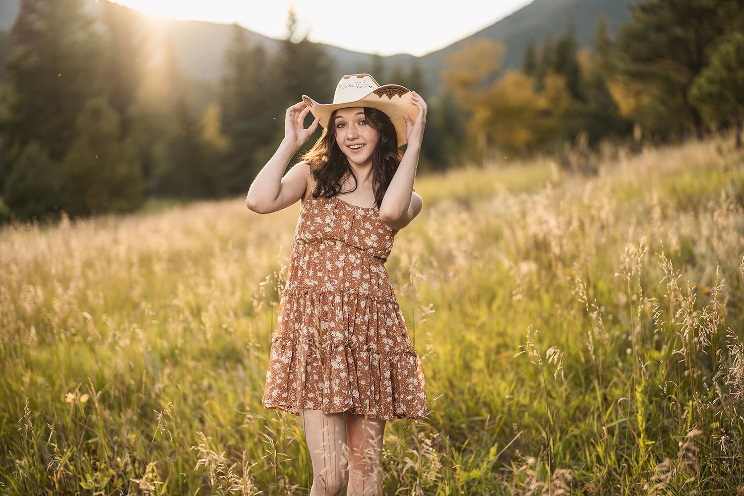 Young woman in floral dress and cowboy hat posing in golden sunlit mountain meadow at sunset.