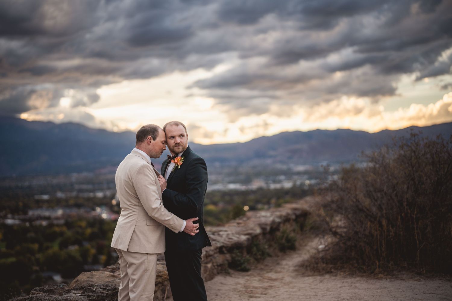 Two grooms embrace on a hilltop at sunset with dramatic clouds and mountain views behind them.