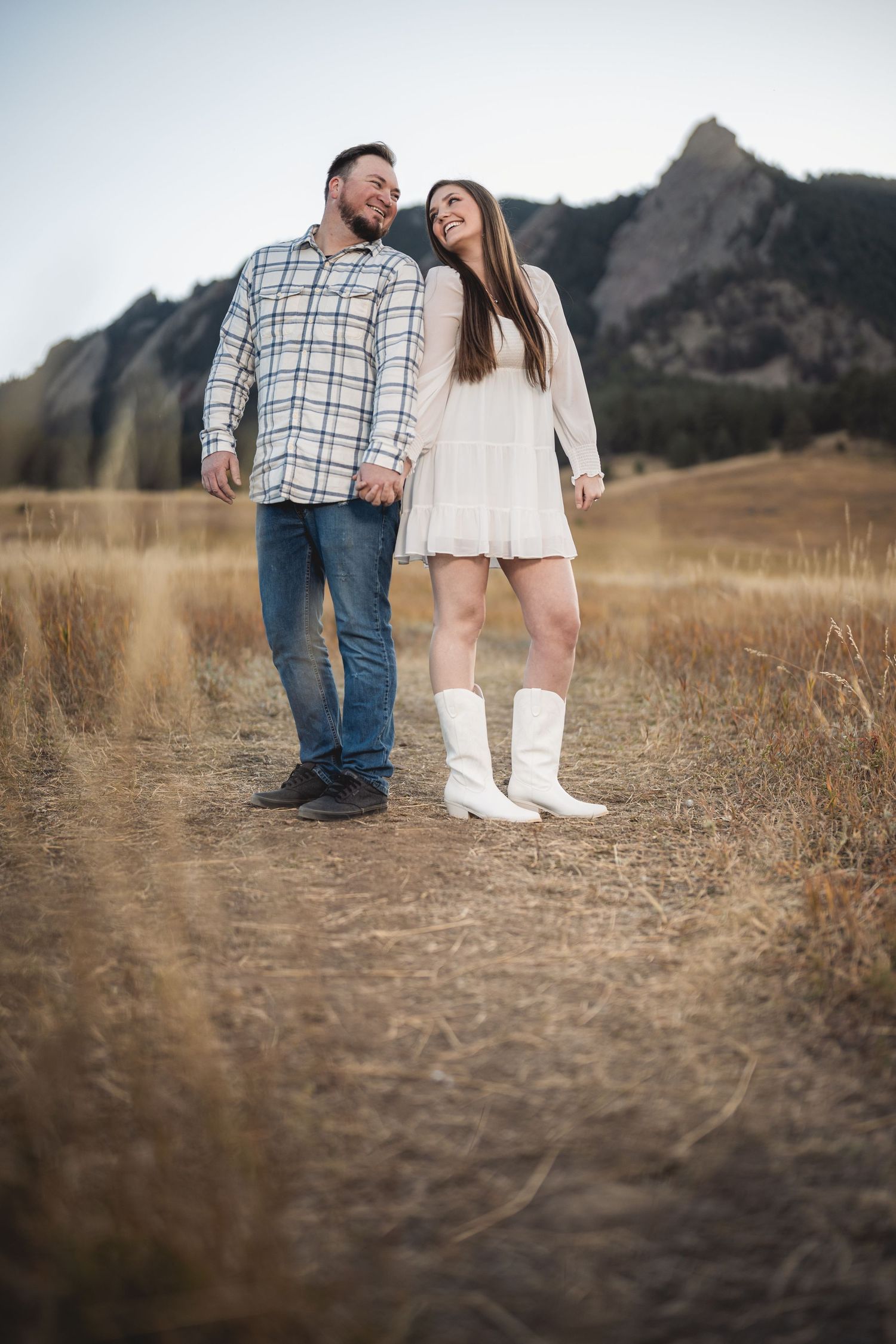 Couple in casual attire stands together in a field with rocky mountain landscape in background.