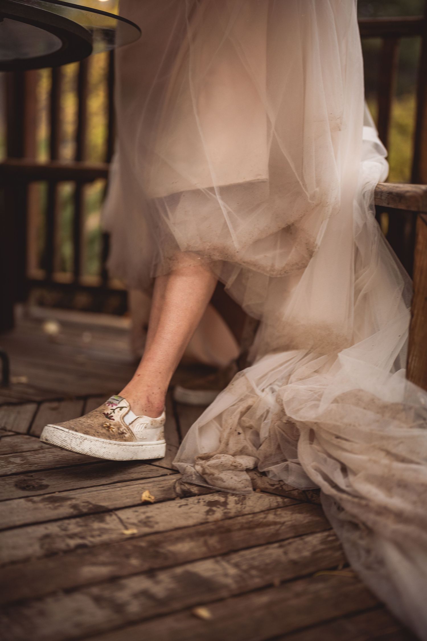Gold sparkly sneakers peek out from under a flowing white wedding dress on a wooden deck.