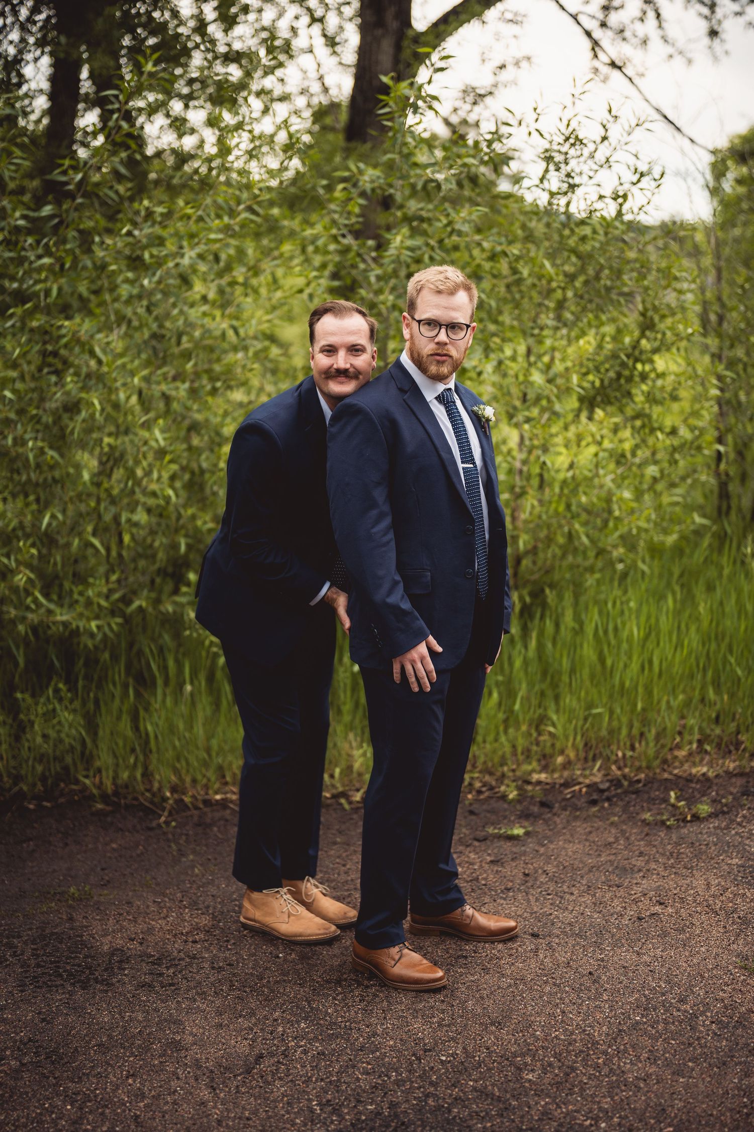 Two people in matching navy suits crouching and laughing together outdoors.