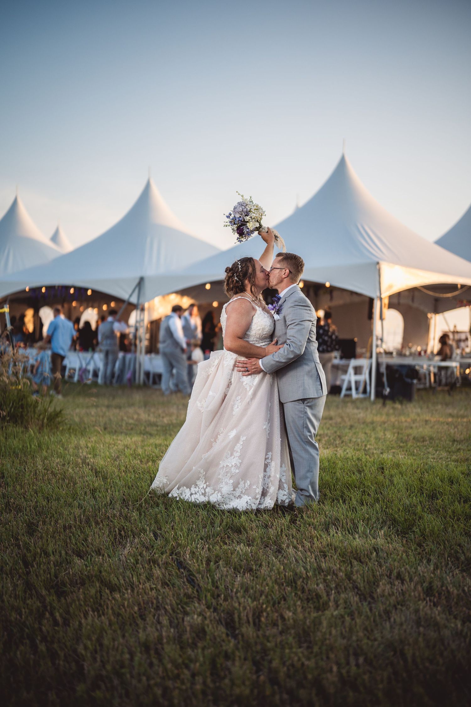 Couple embraces on grassy field with white reception tents in background during sunset.