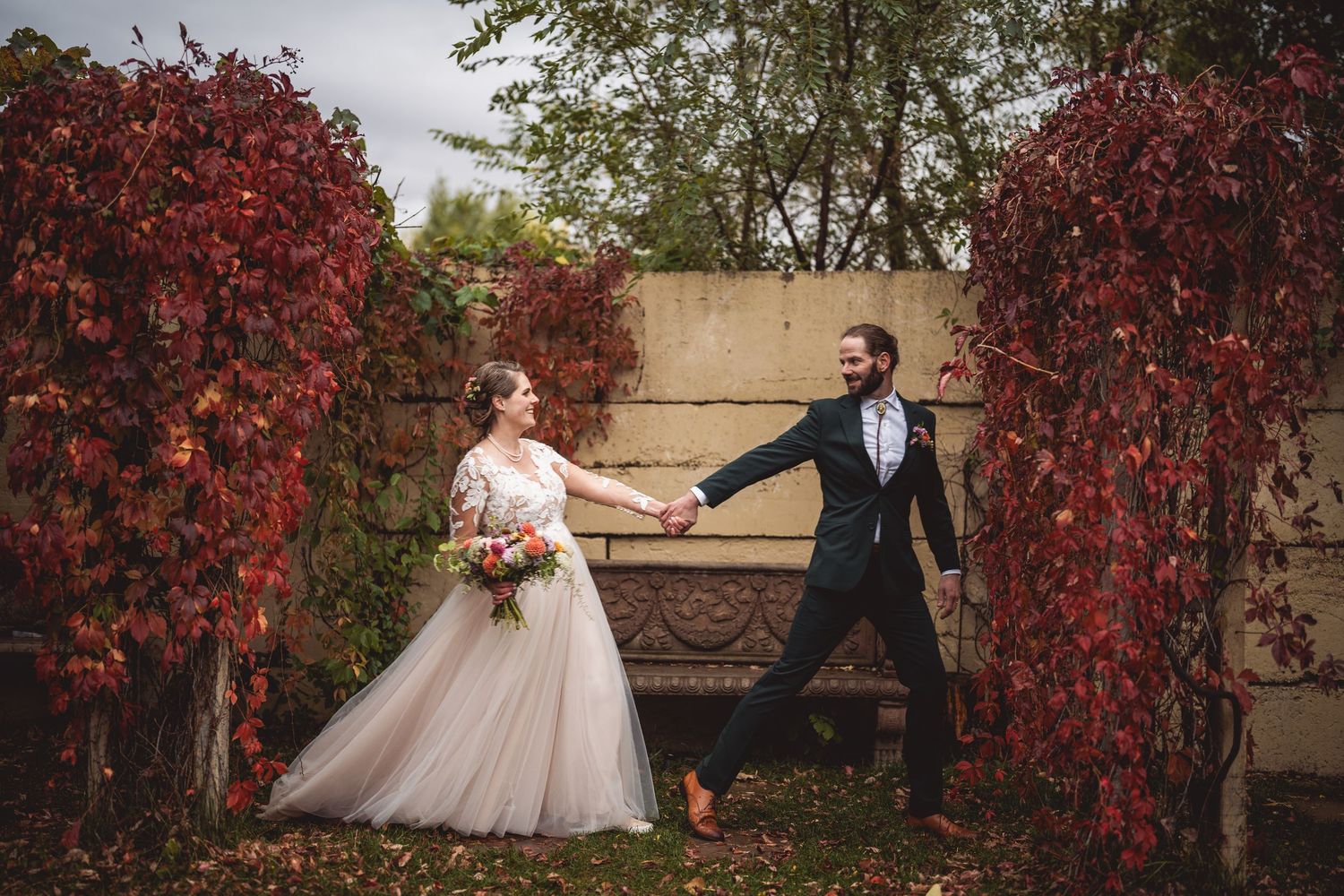 Playful wedding couple dancing between red vine-covered walls in garden setting.