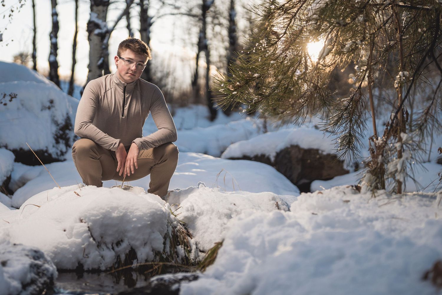 Man in beige outfit sitting on snow-covered rocks in a winter forest with sunlight filtering through pine trees.