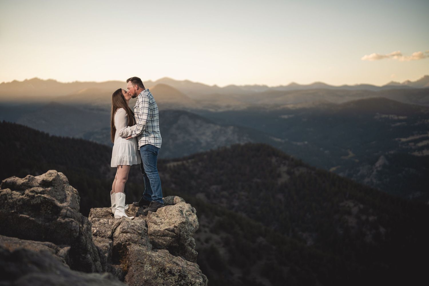 Couple kissing on rocky mountain summit at sunset with scenic valley and mountain range backdrop.