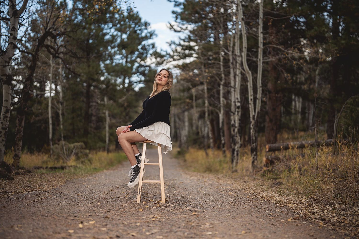 Smiling blonde woman sitting on stool on forest dirt road in black outfit.