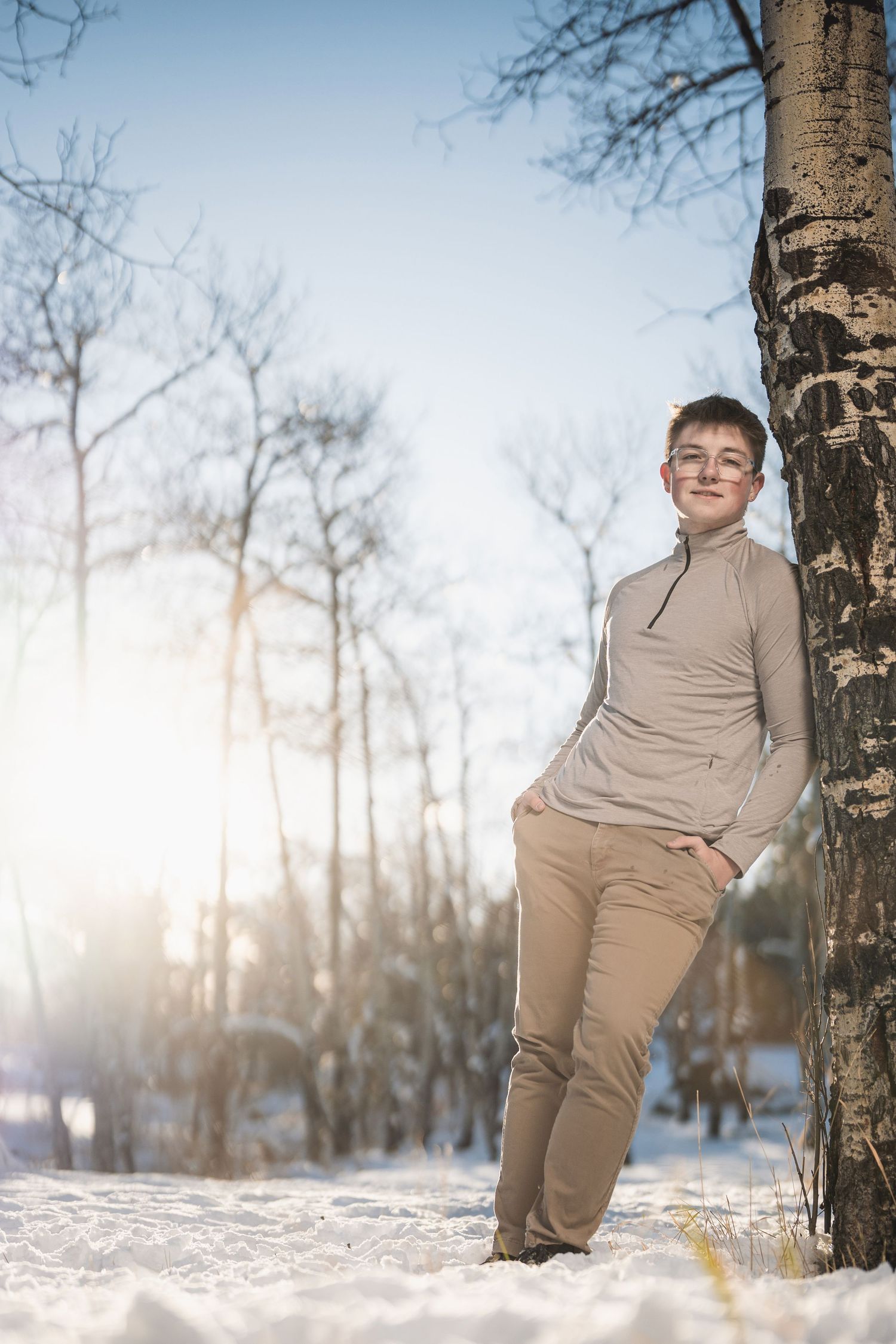 Teen boy in glasses leans against birch tree in snowy winter park wearing beige turtleneck and khakis.