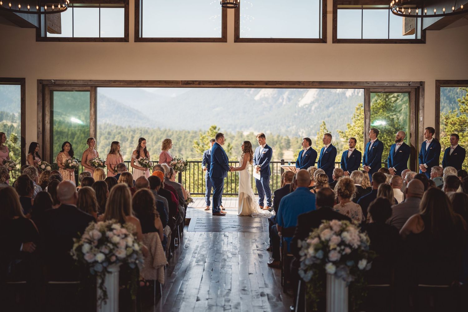 Indoor wedding ceremony with mountain views, couple exchanging vows at altar with wedding party and guests seated in rows.