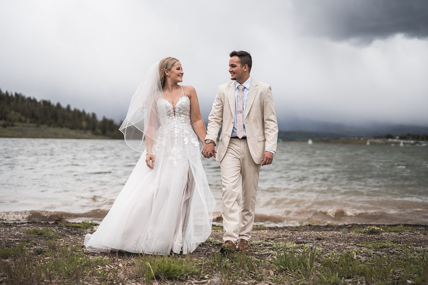 Bride and groom walking hand in hand by a mountain lake on their wedding day, wearing white gown and tan suit.