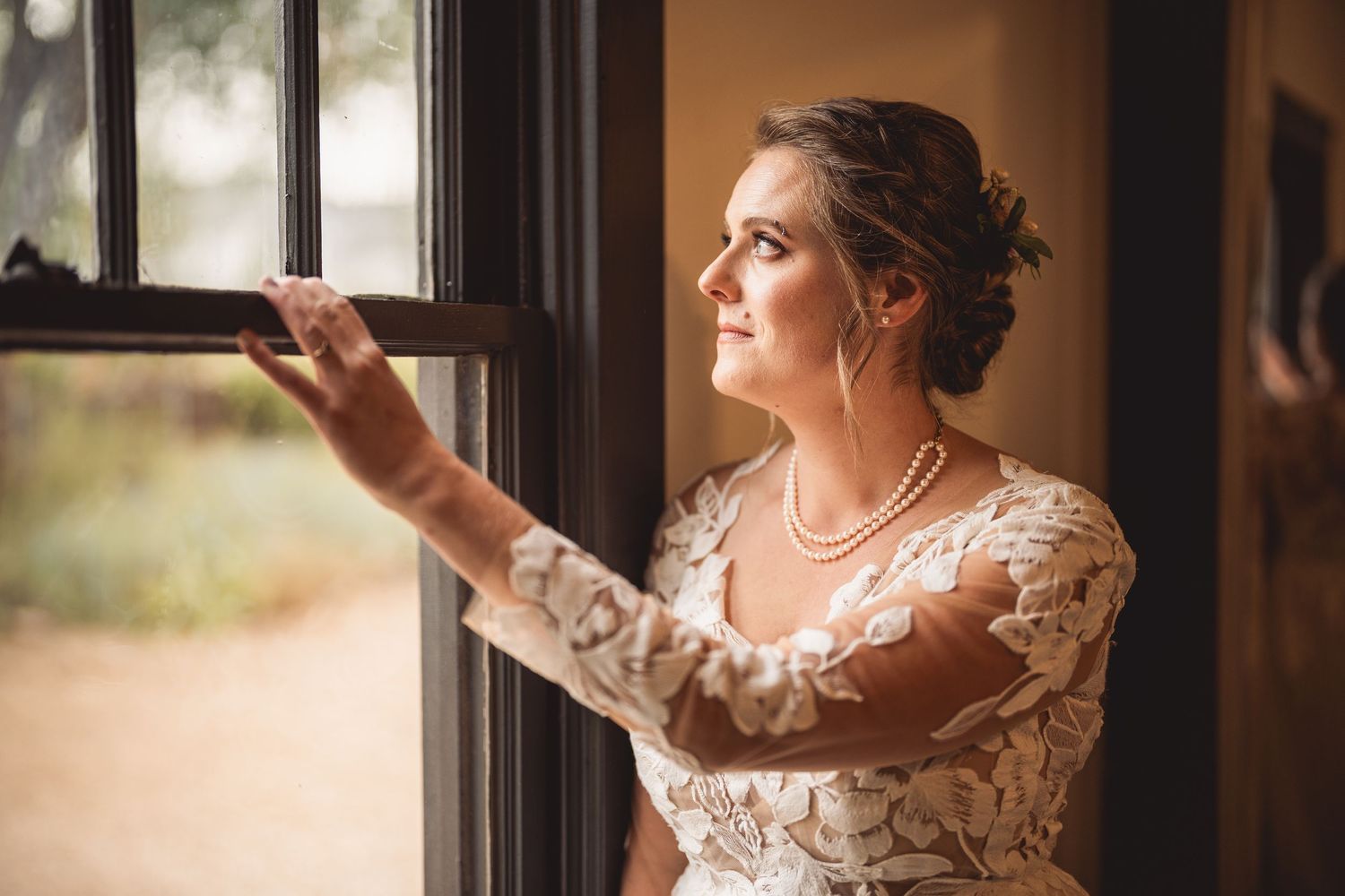 Bride in lace wedding dress gazes out a rustic window, wearing pearl necklace with elegant updo hairstyle.
