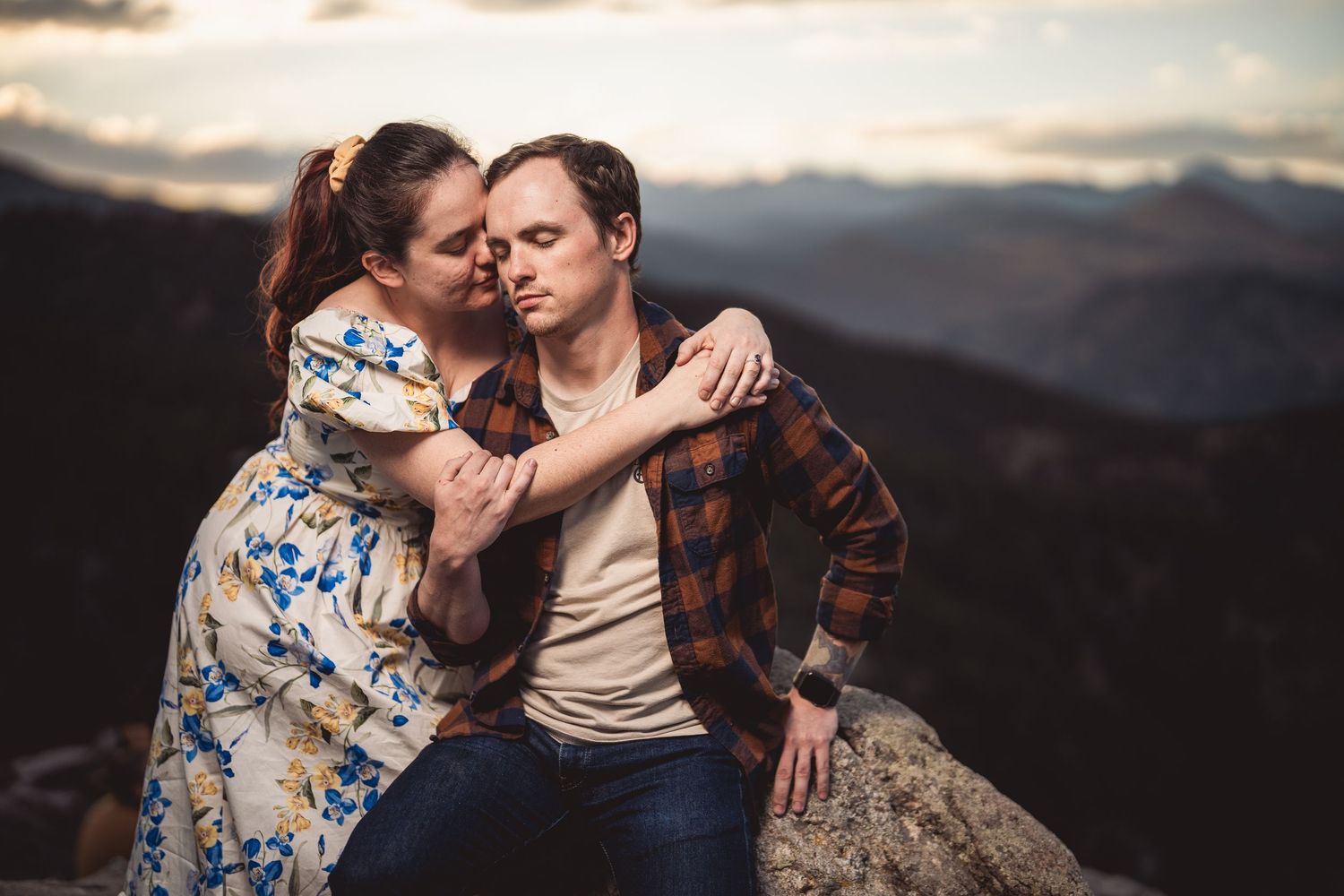 Couple embracing on mountain rock at sunset, woman in floral dress hugging man in plaid shirt with scenic valley backdrop.