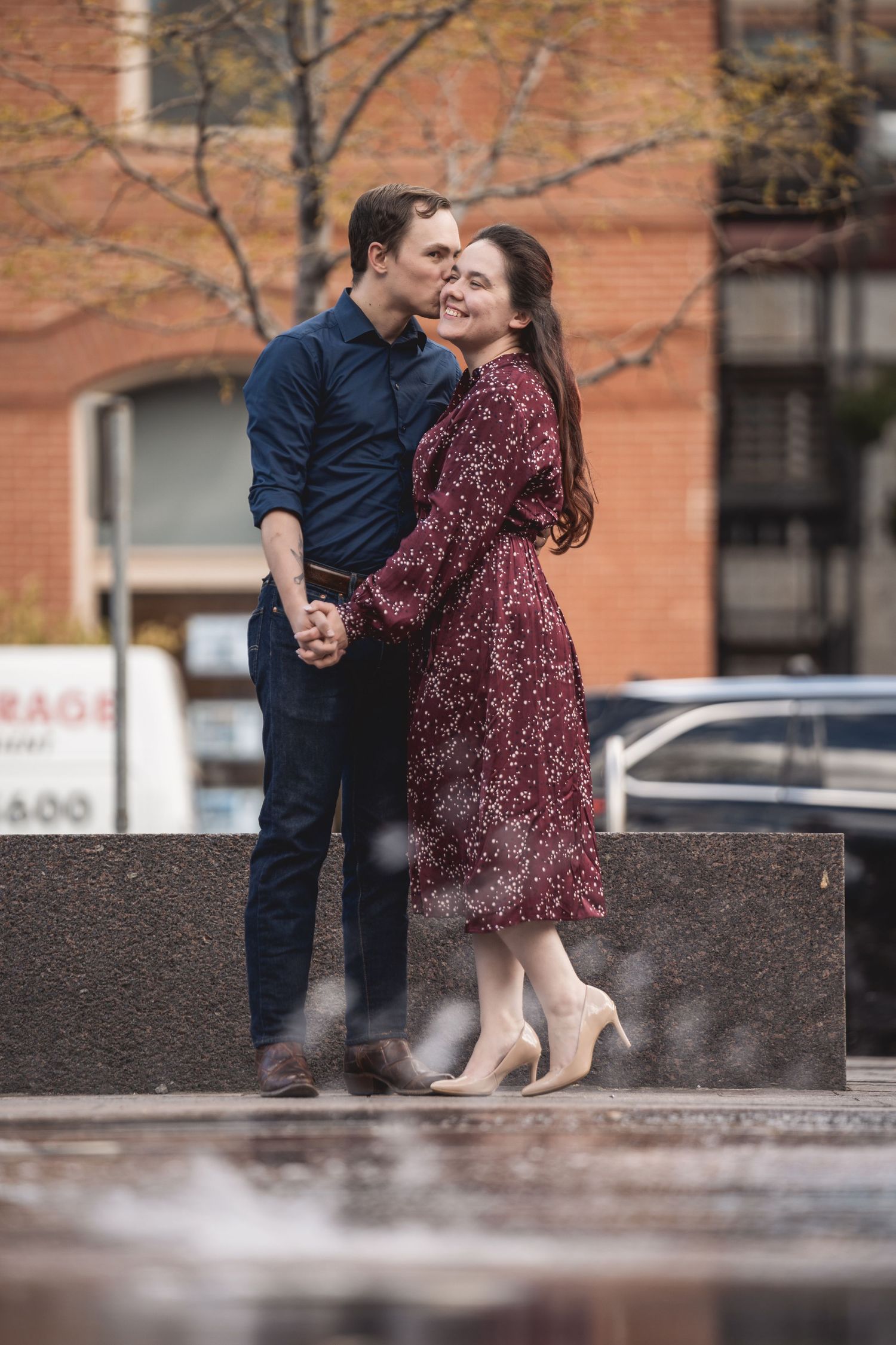 Couple embracing and kissing outdoors on a rainy day, woman in burgundy floral dress and man in navy blue shirt.