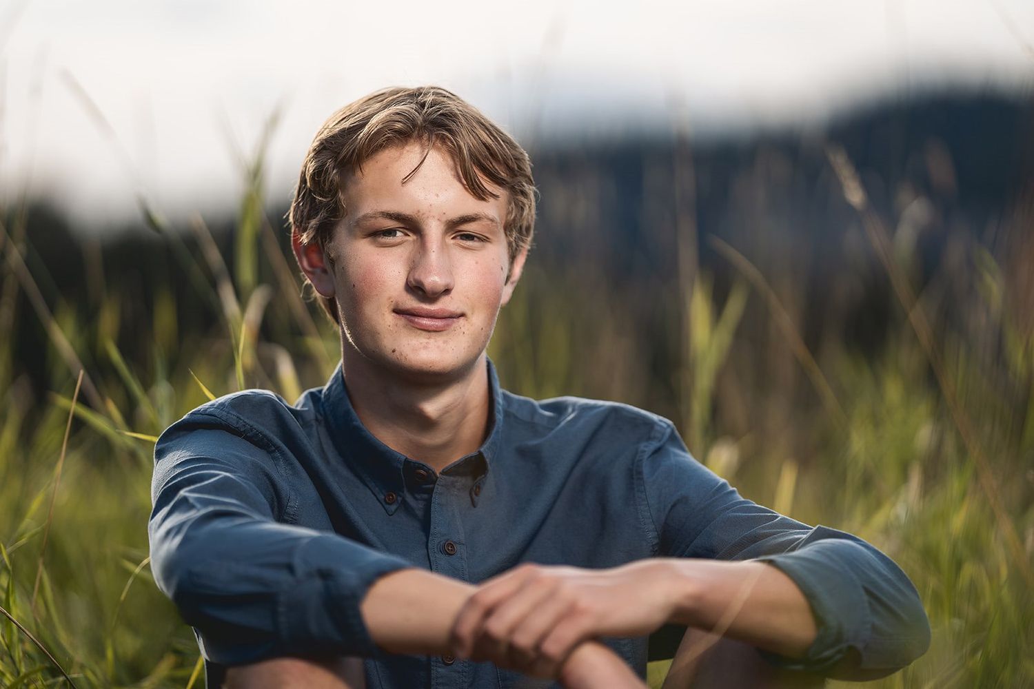 Casual senior portrait of someone in a blue shirt sitting in tall grass at sunset.