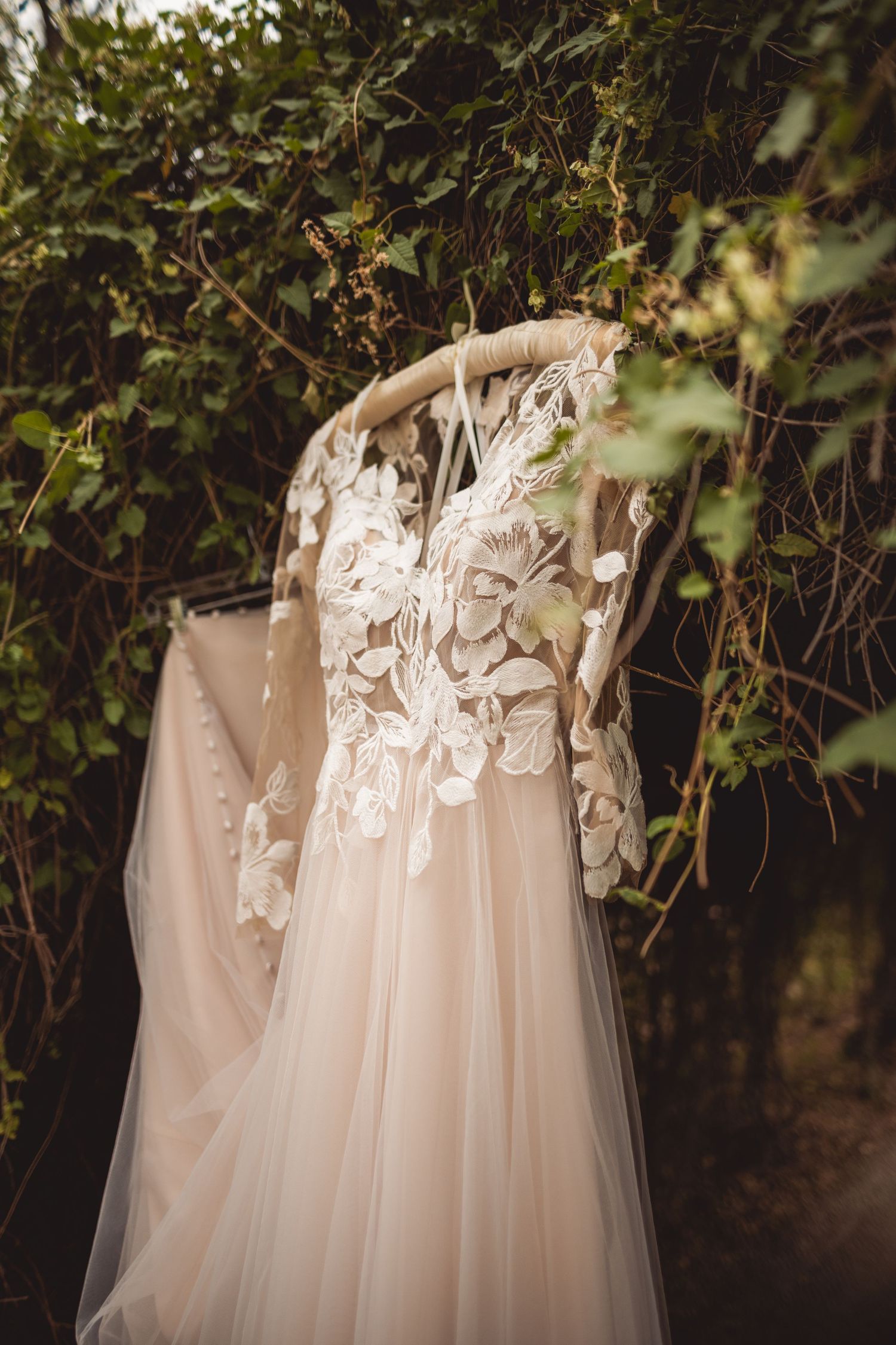 Elegant white wedding dress with lace details hanging among green garden foliage.