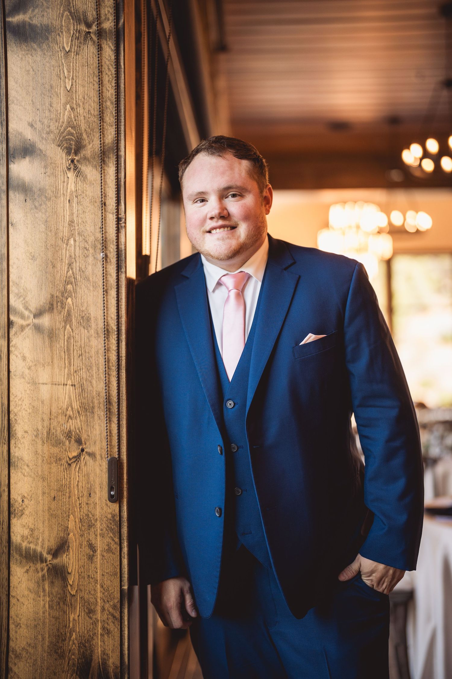 Person in navy three-piece suit with pink tie standing by wooden column.
