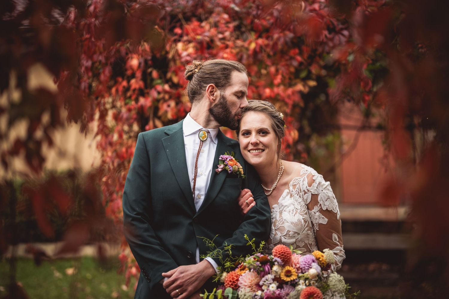 Wedding portrait featuring couple beneath red fall leaves holding colorful autumn bouquet.
