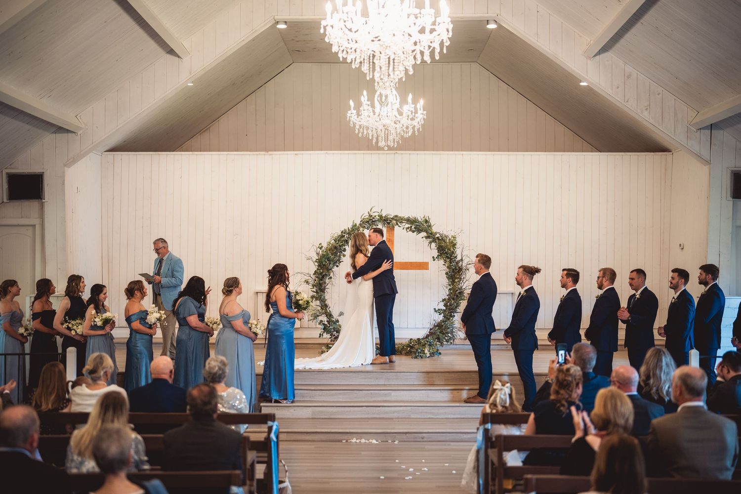 Wedding ceremony in white chapel with floral arch and rows of guests in blue attire.