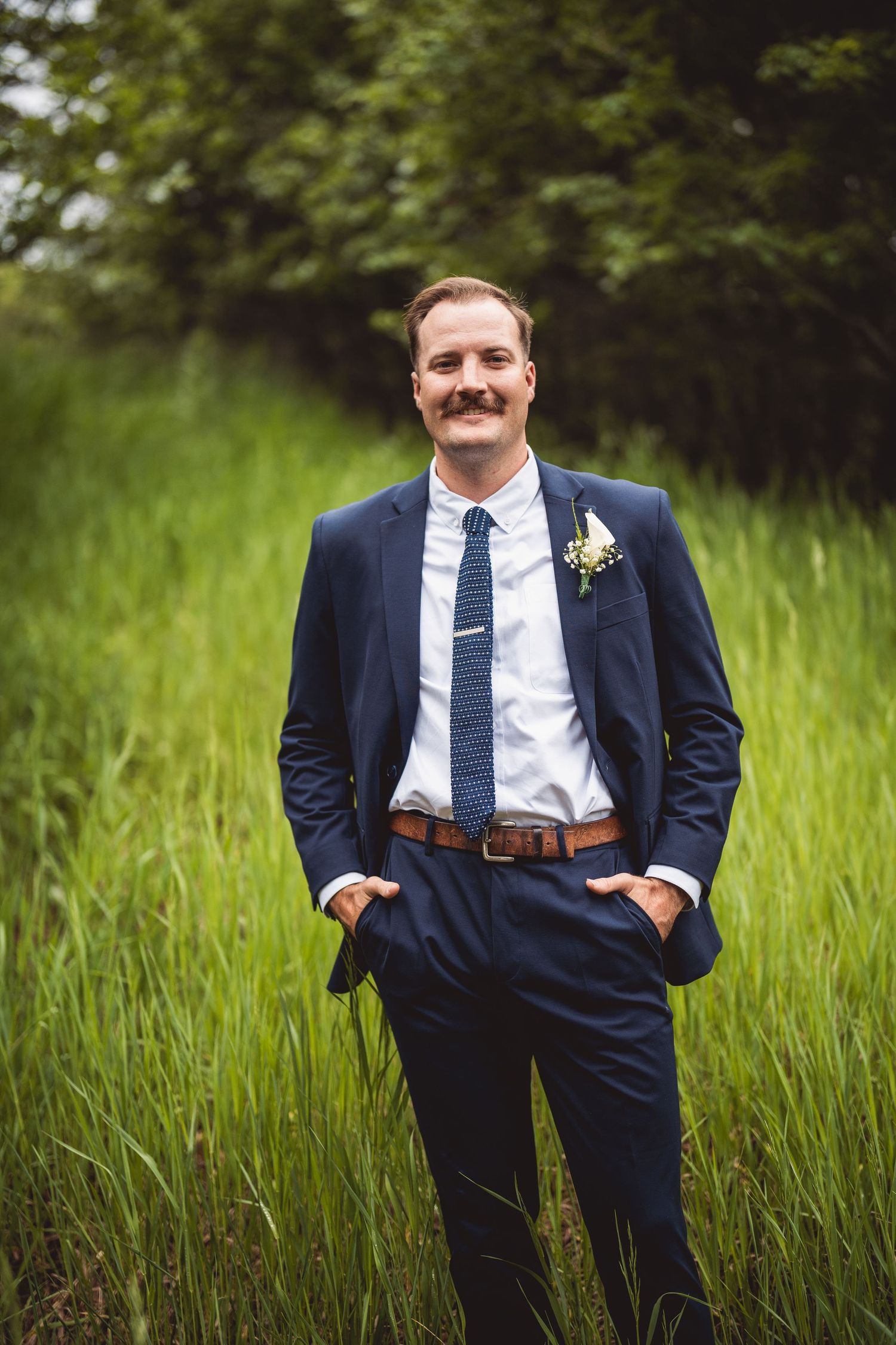 Wedding portraits of a groom in a navy suit and tie standing in tall grass.