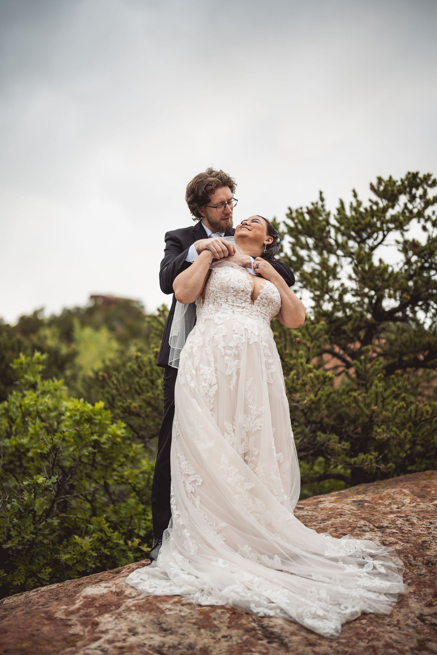 A romantic wedding couple embraces on a rocky overlook with a flowing white lace dress trailing behind them.