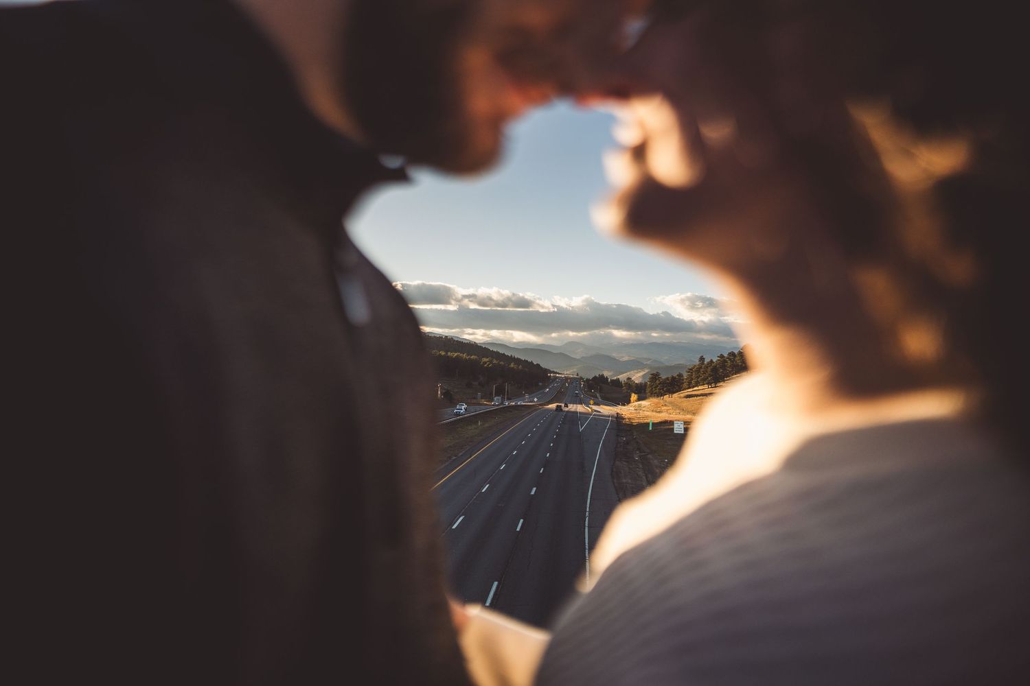 Romantic couple sharing a kiss at golden hour with a scenic highway and mountain landscape in the background.