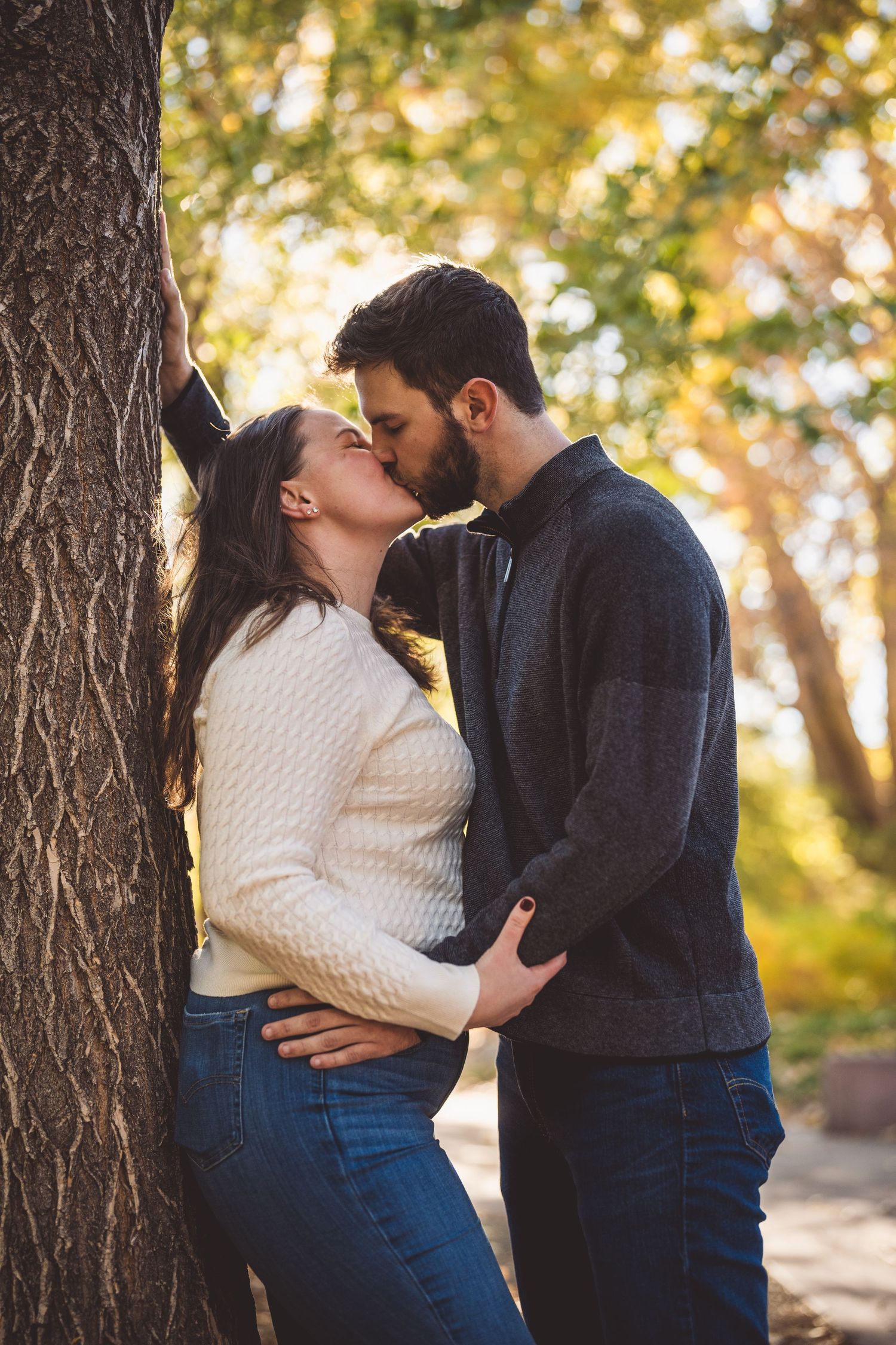 A couple shares a romantic kiss leaning against a tree in a sunlit autumn park, wearing casual sweaters and jeans.