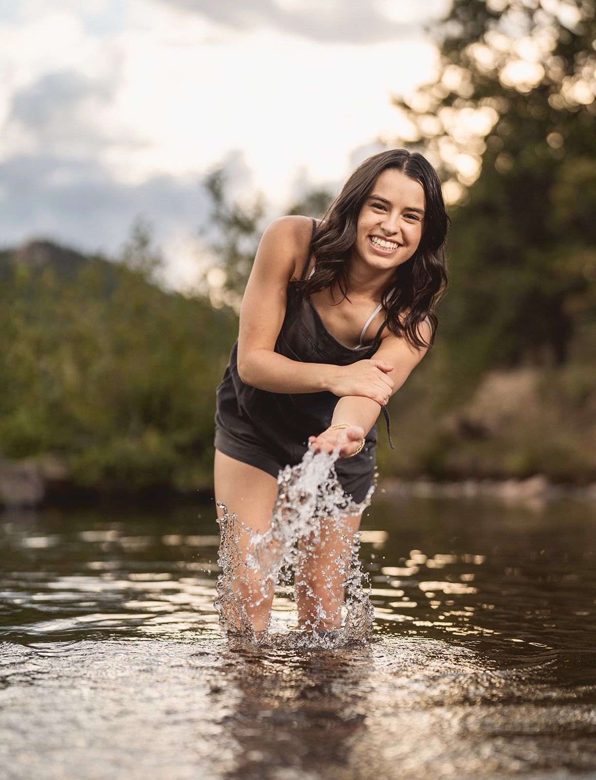 Young woman laughing and splashing water in a shallow river, wearing a black swimsuit, with trees and sky in background.