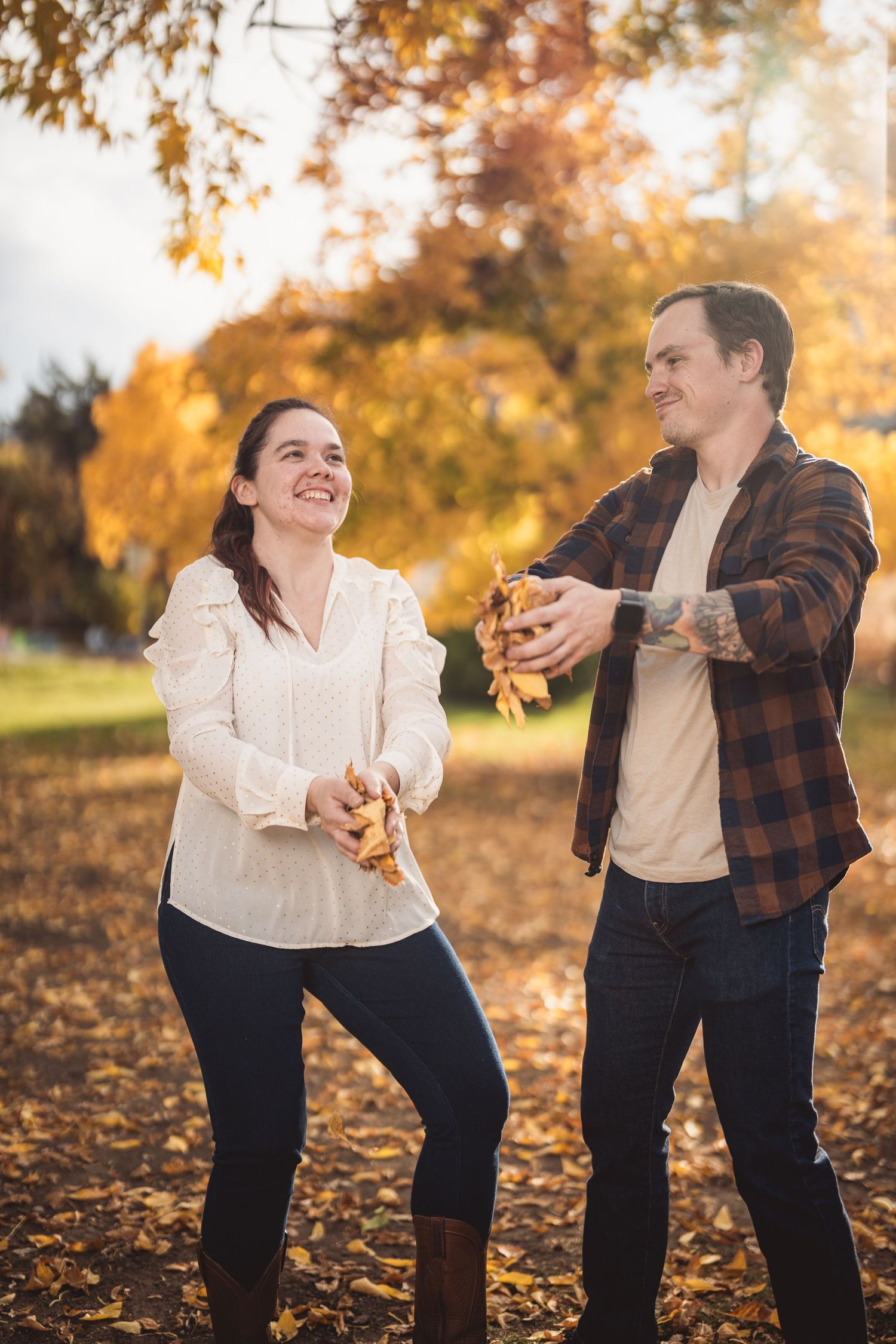 Couple laughing and tossing autumn leaves in a sunlit park with golden fall foliage in the background.