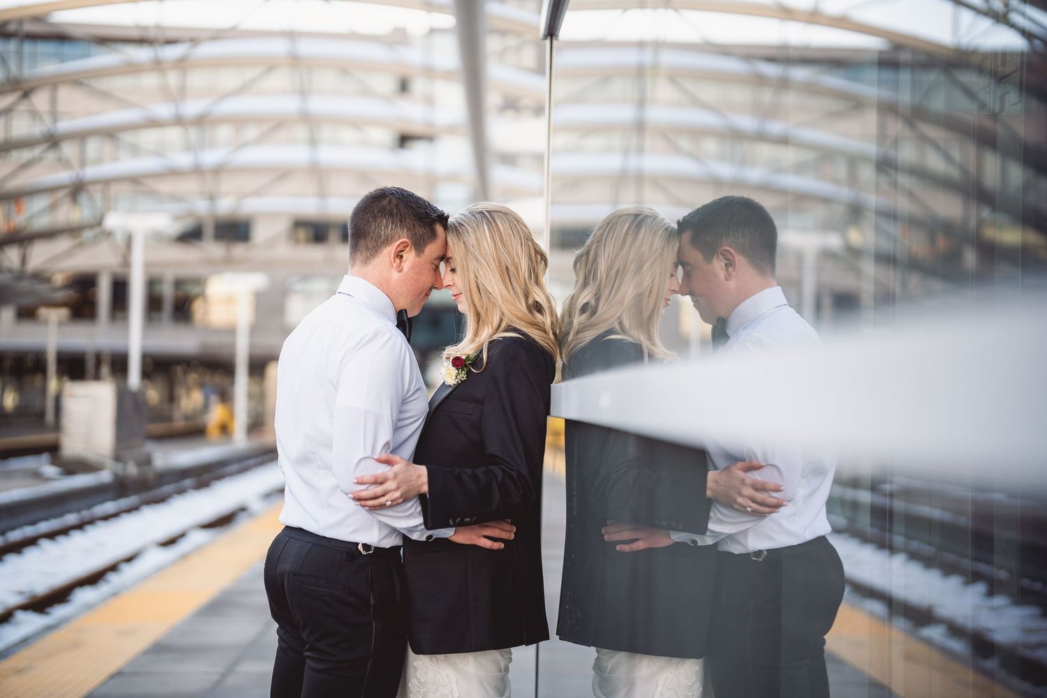 Couple sharing an intimate moment on a train station platform, with their reflection visible in a glass window.