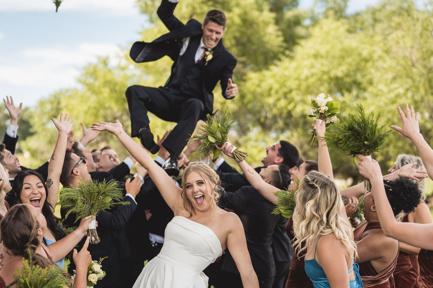 Joyful wedding party tosses groom in air as bride smiles in white dress, surrounded by bridesmaids outdoors.