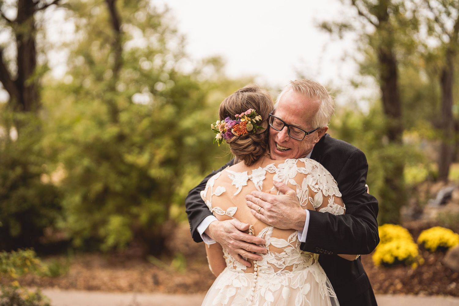 Father embracing bride in lace wedding dress with floral hair wreath outdoors at wedding ceremony.