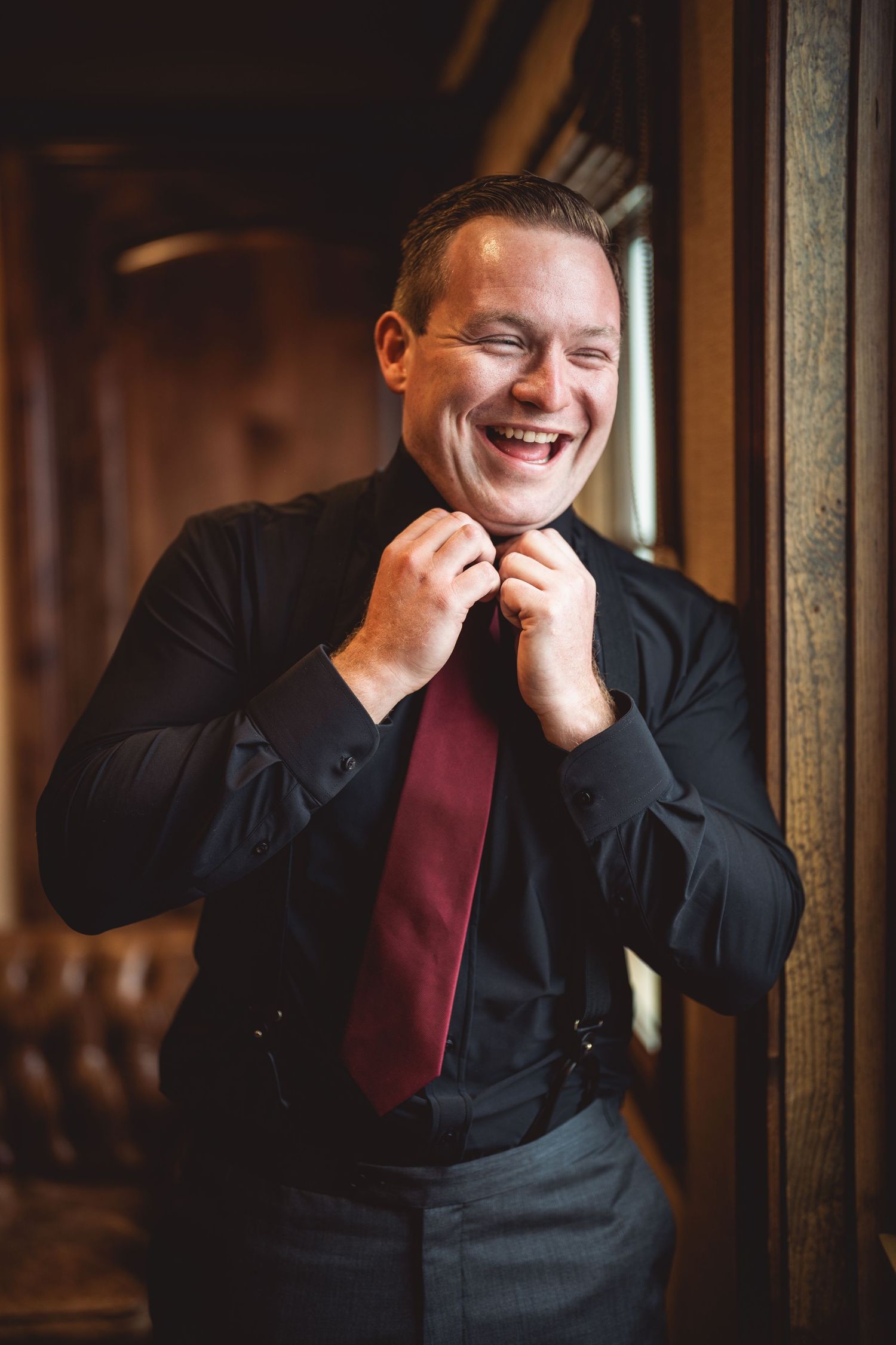 A person adjusts their burgundy tie while laughing joyfully in an indoor setting.