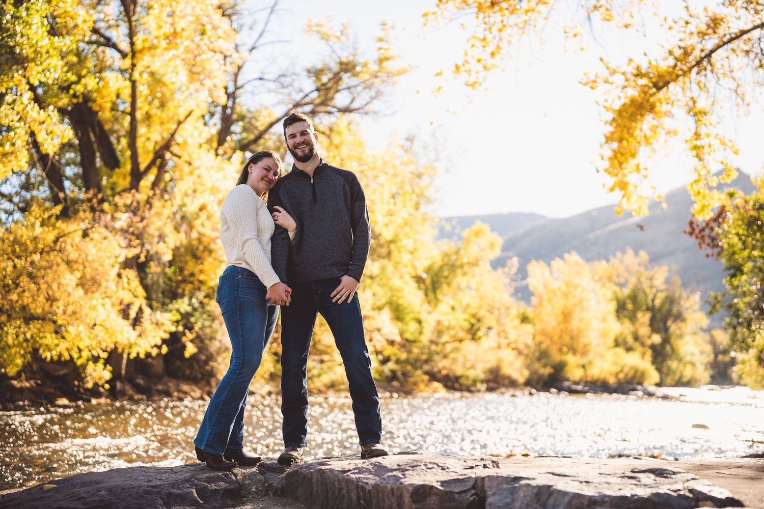 A couple poses together on natural terrain with golden autumn trees in the background.