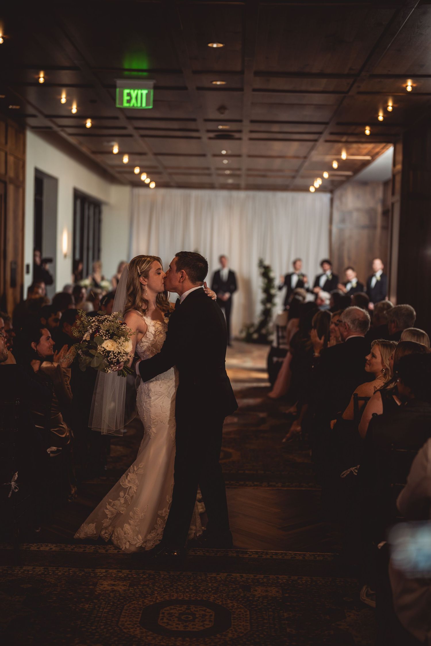 A dimly lit indoor wedding ceremony with guests seated in rows and ambient lighting.