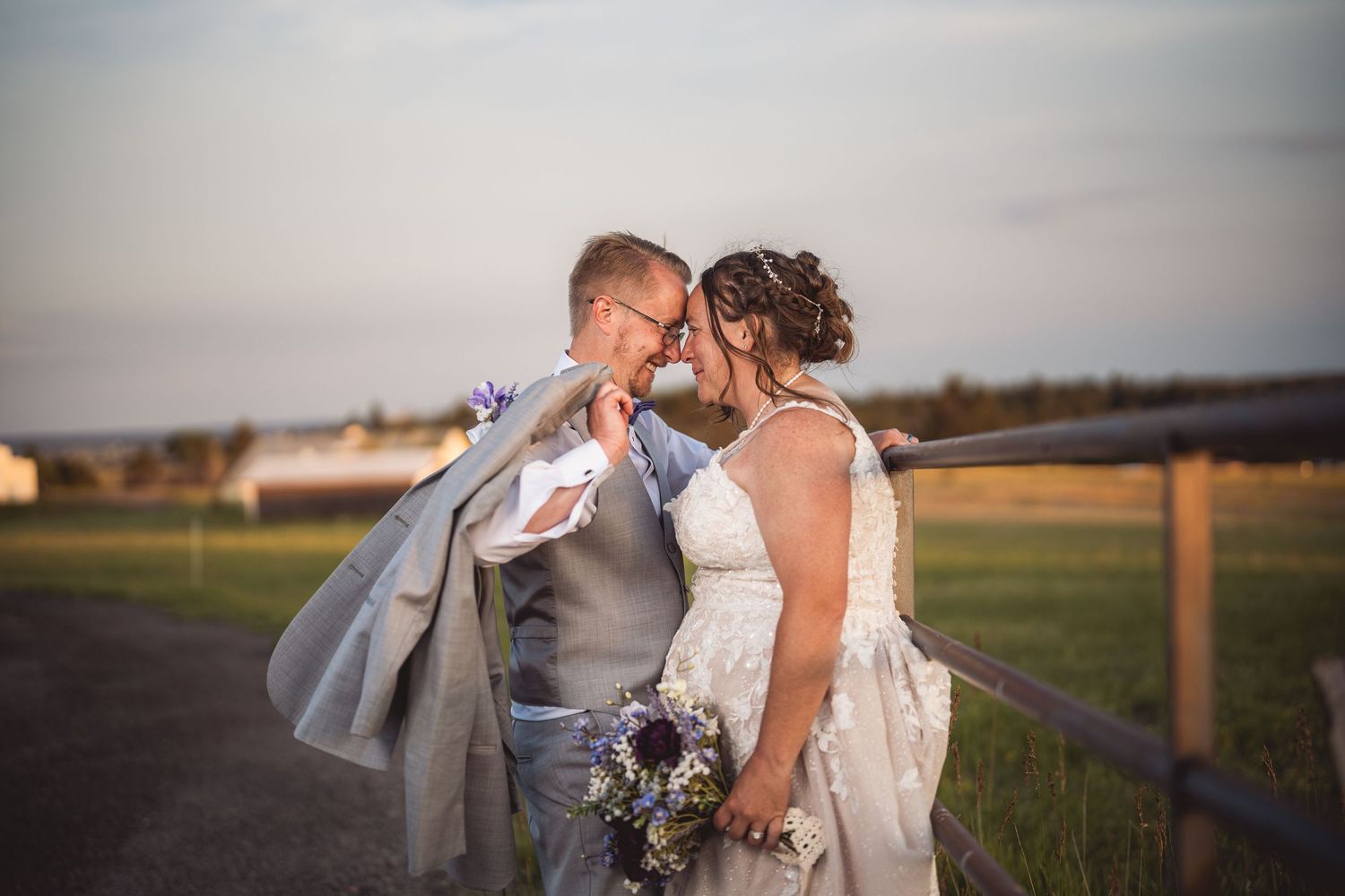 Bride and groom share a romantic moment at sunset by a rustic fence in a scenic countryside wedding setting.
