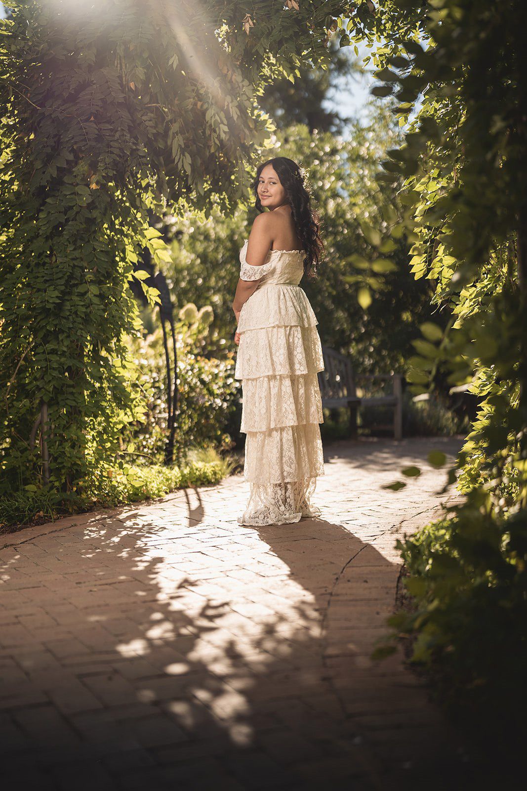Woman in white off-shoulder lace gown standing on brick path surrounded by lush green garden foliage in sunlight.