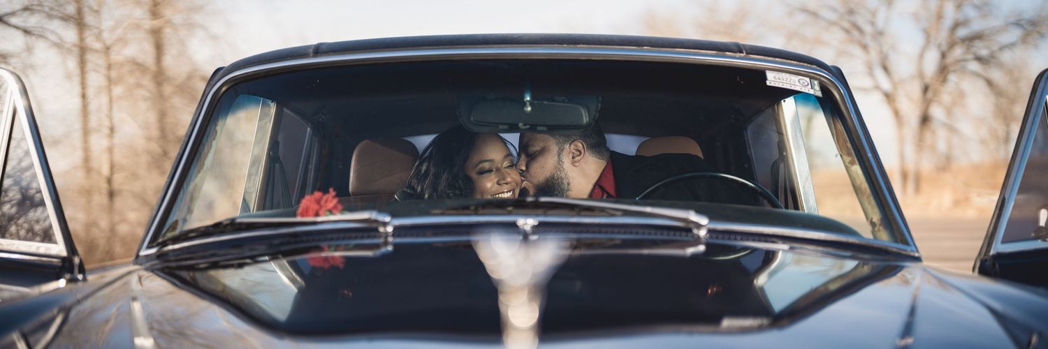 A couple shares a romantic kiss inside a vintage black car, viewed through the windshield on a soft autumn day.