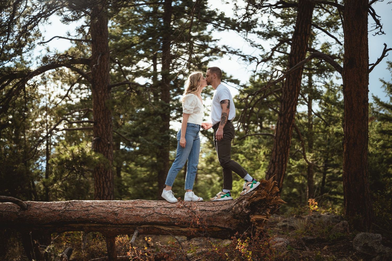 A romantic couple sharing a kiss while balancing on a fallen log in a lush pine forest.