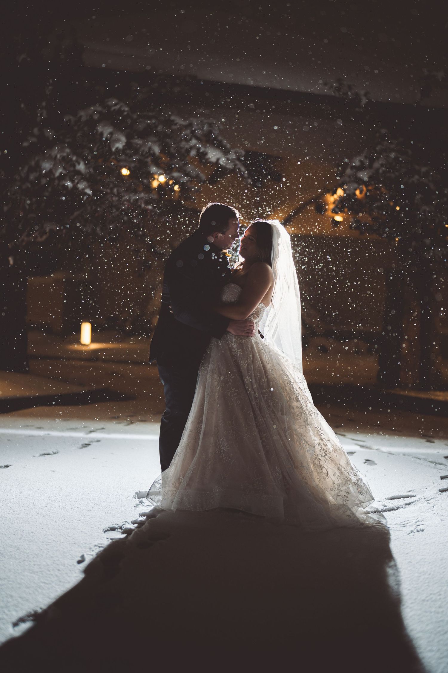 Nighttime winter wedding portrait with couple silhouetted against snowy backdrop.