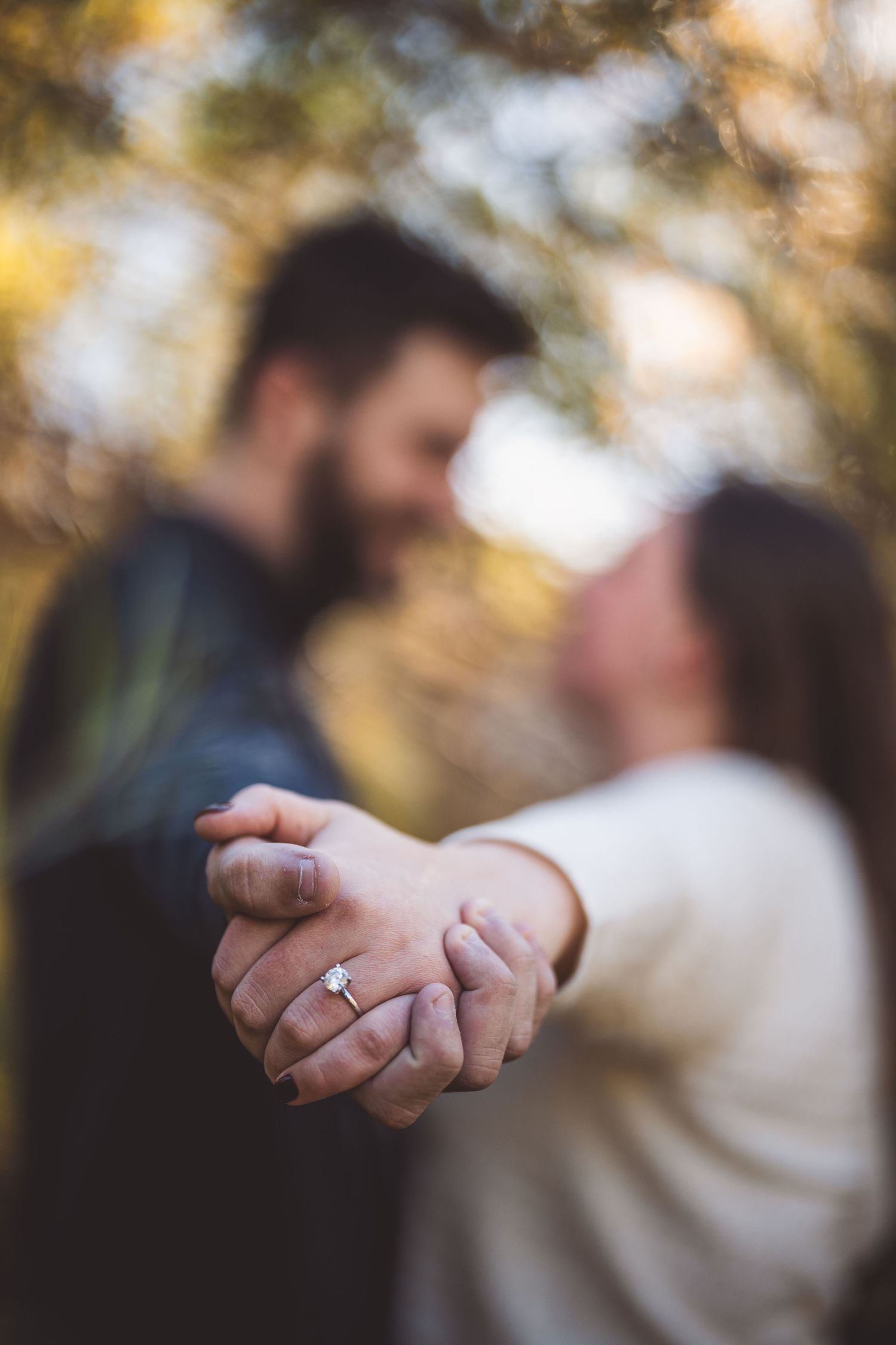 Close-up detail shot of hands and engagement ring with soft autumn bokeh.