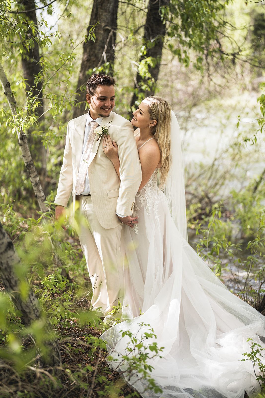A bride and groom share a romantic moment in a lush green forest, she in a white gown, he in a beige suit.